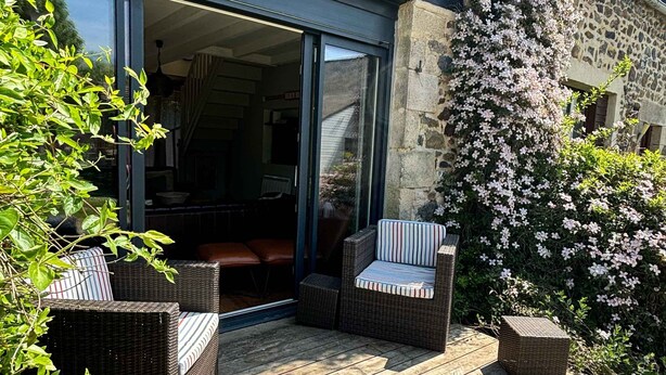 Een gebouw van steen met een houten terras, rieten meubels en bloeiende planten.