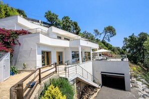 Une maison moderne à deux étages, dotée d’un balcon, d’un garage et d’un jardin agrémenté de plantes et de fleurs.