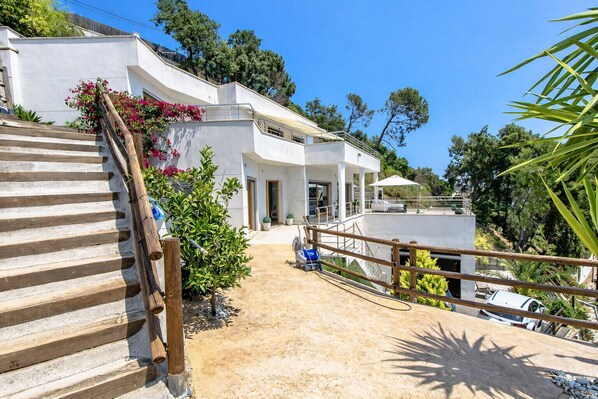 Une villa moderne avec un escalier menant à un espace extérieur couvert, entourée de verdure et surplombant un ciel bleu azur.