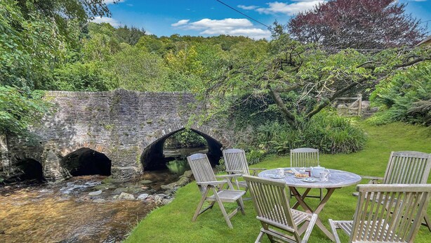 A rustic stone bridge over a stream, surrounded by lush greenery and a wooden table with chairs. - Ball Cottage, Winsford (Winsford)