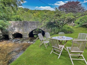 Un pont de pierre rustique enjambant un ruisseau, entouré d’une végétation luxuriante, avec une table en bois et des chaises à proximité.