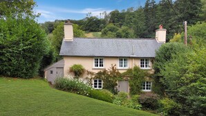 A house with a sloped roof, multiple chimneys, and a garden with various plants and flowers. - Ball Cottage, Winsford (Winsford)