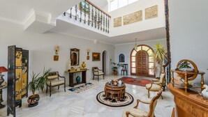 Un interior espacioso y luminoso, con techo alto, una imponente escalera y diversos muebles, incluyendo una gran mesa redonda, sillas y un aparador decorativo.