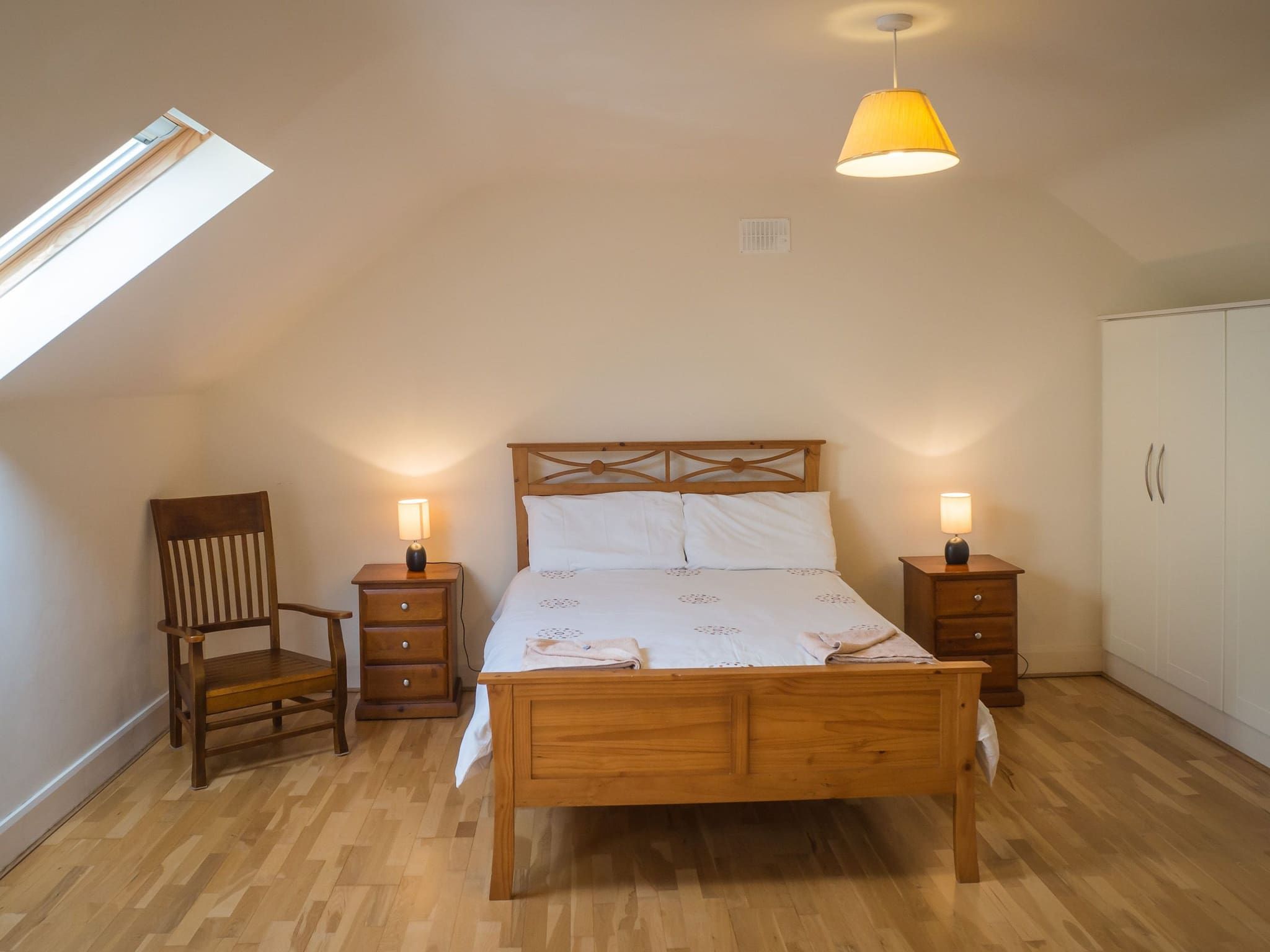 Ein Schlafzimmer mit einem hölzernen Bett, zwei Nachttischen mit Lampen, einem Holzstuhl und einem Kleiderschrank.