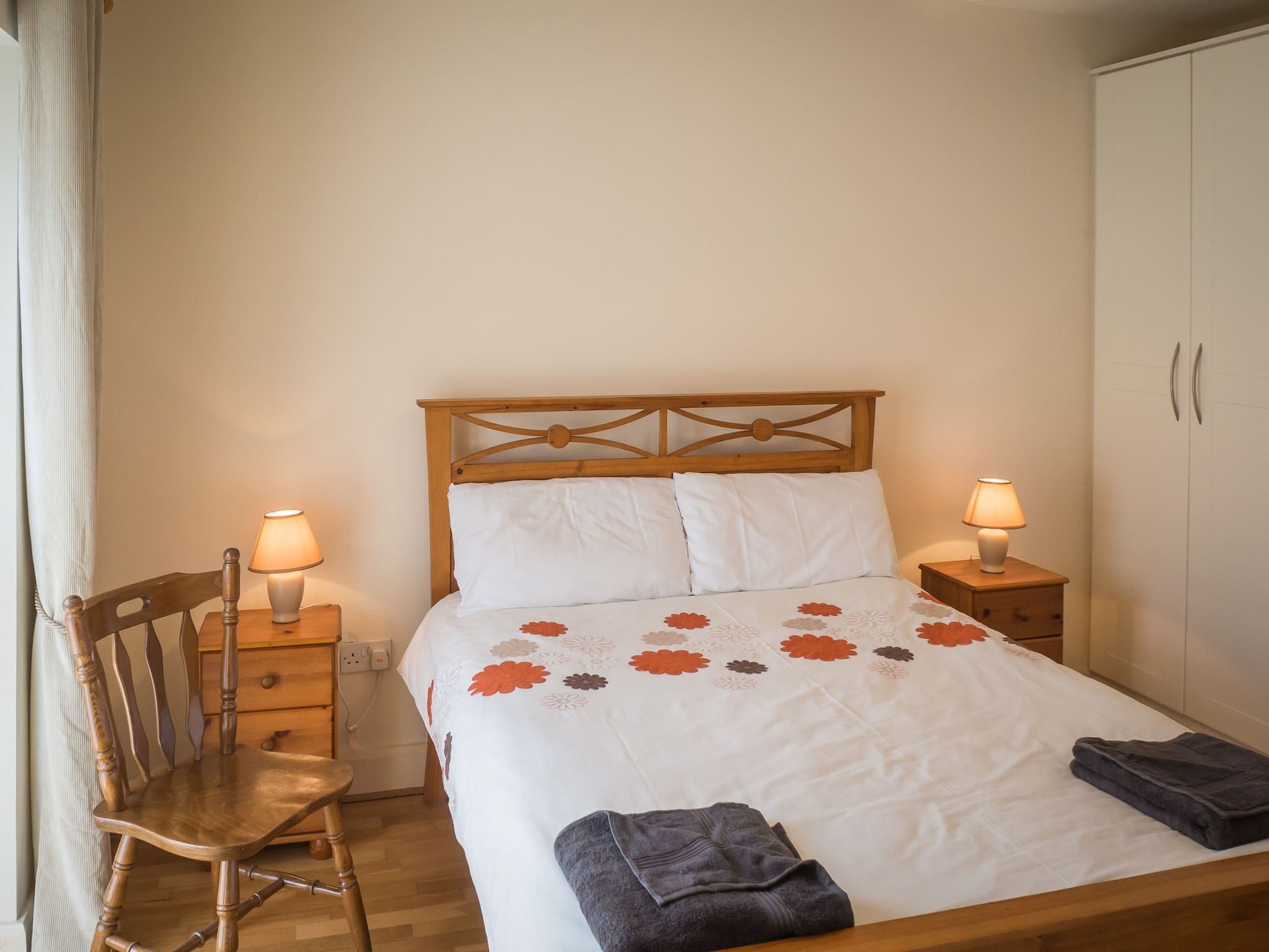 A neatly made bed with a floral bedspread, a wooden nightstand with a lamp, a wooden chair, and a wardrobe.