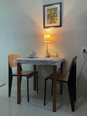 Een eettafel met een kant kleed, twee stoelen, een tafellamp, een ingelijst beeld en een stopcontact aan de muur.