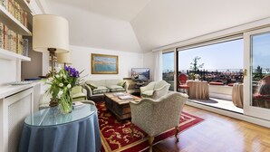 A living room with a sofa, armchairs, a coffee table, and a glass table with a vase of flowers. There is a bookshelf with books and a painting on the wall. The room has a large window with a view of a cityscape. - Ca' Cerchieri Terrace Canal View (Venice)