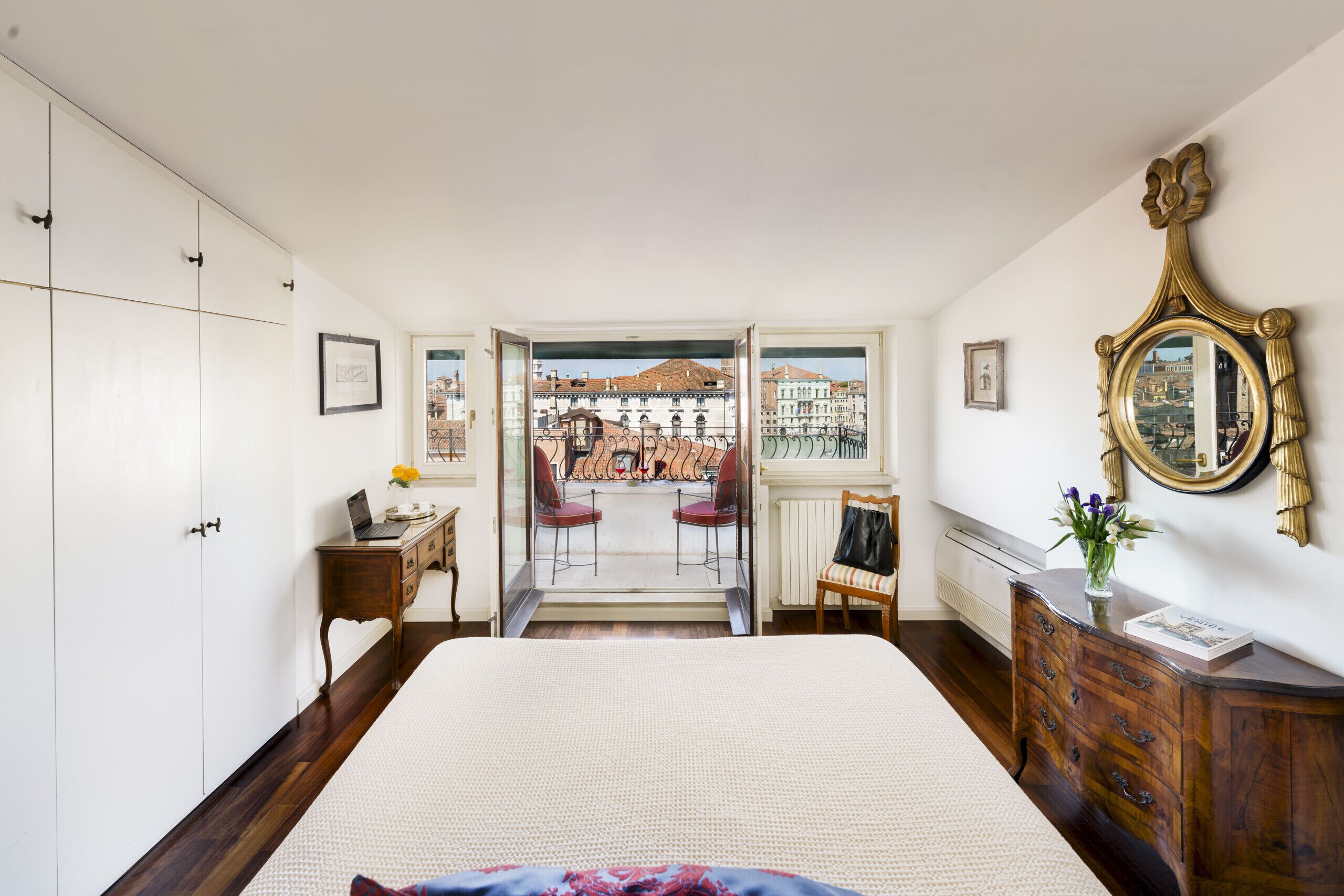A bedroom with a bed, a desk, a chair, a dresser, and a large window with a view of buildings.