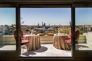 Une terrasse avec deux tables et des chaises, une vue sur un paysage urbain avec des bâtiments et une tour, et un ciel dégagé.