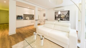 A modern living room with a white sofa, a glass coffee table, and a kitchen in the background.