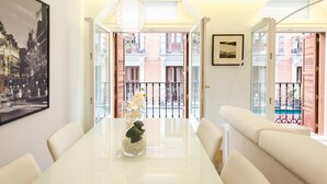 A dining area with a white table, chairs, a vase with flowers, a pendant light, and a framed picture on the wall.