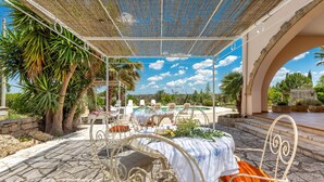 An outdoor dining area with a table and chairs under a pergola, overlooking a pool and garden.