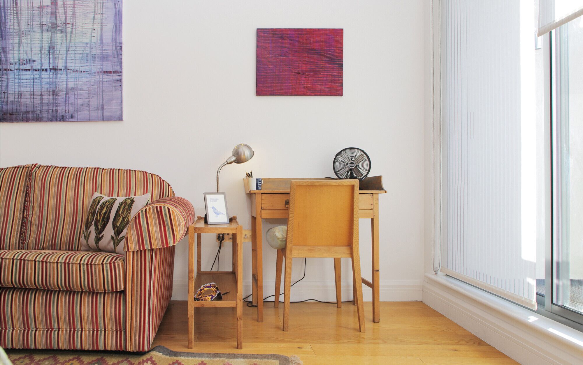 A living room with a striped sofa, a wooden desk, and a chair. There is a lamp on the desk and a picture frame.