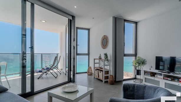 Ein modernes Wohnzimmer mit Balkon und Meerblick, inklusive Fernseher und Bücherregal.