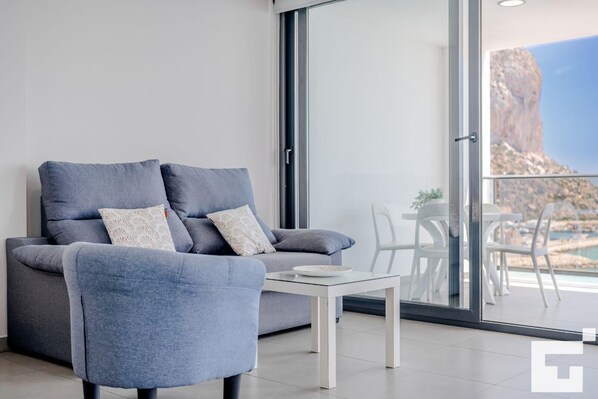 A modern living room with a grey sofa and armchair, a small white table, and a large sliding glass door leading to an outdoor terrace with a view of a mountainous landscape.