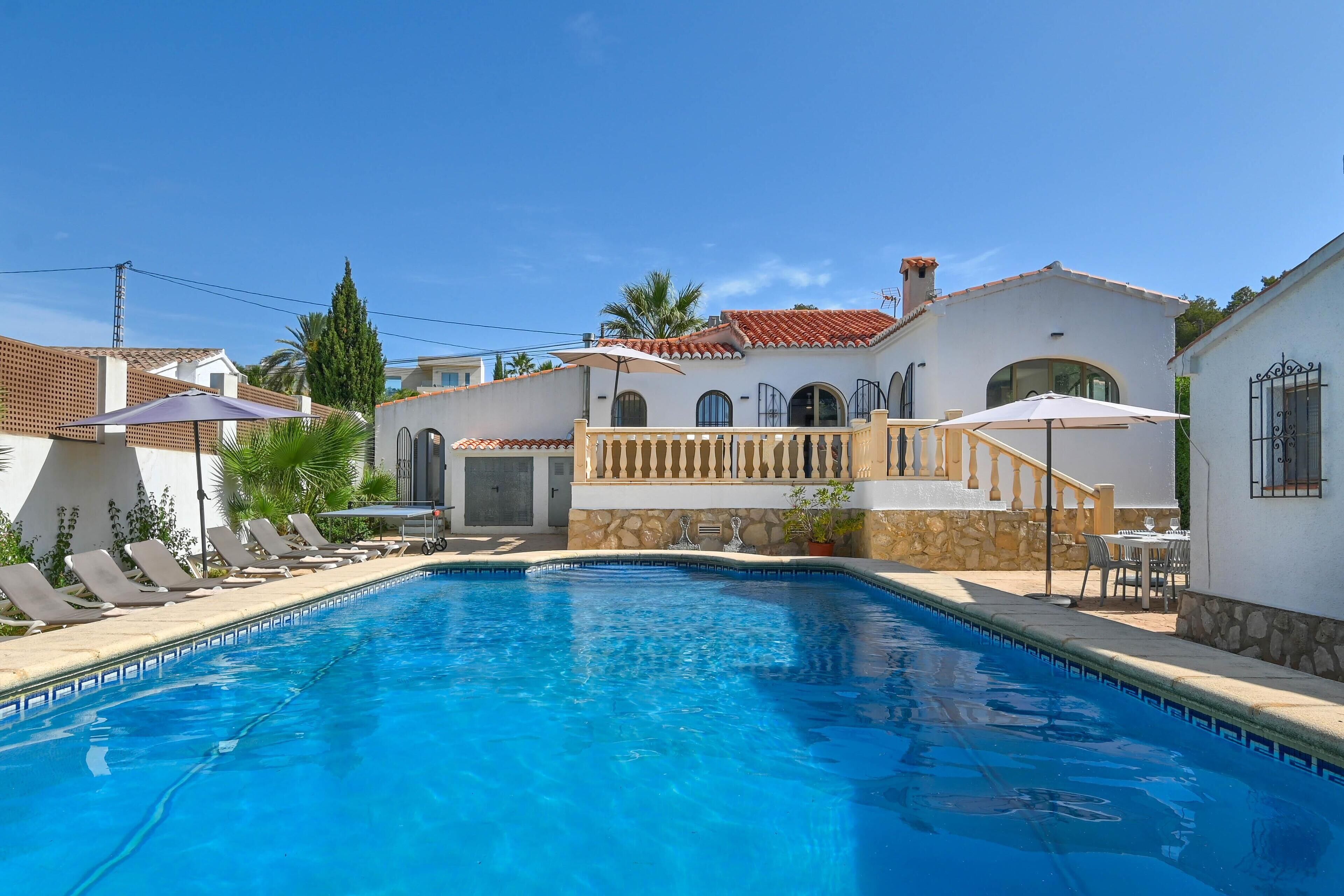 Une piscine avec des chaises longues, un mur en pierre et un bâtiment blanc surmonté d’un toit recouvert de tuiles rouges.