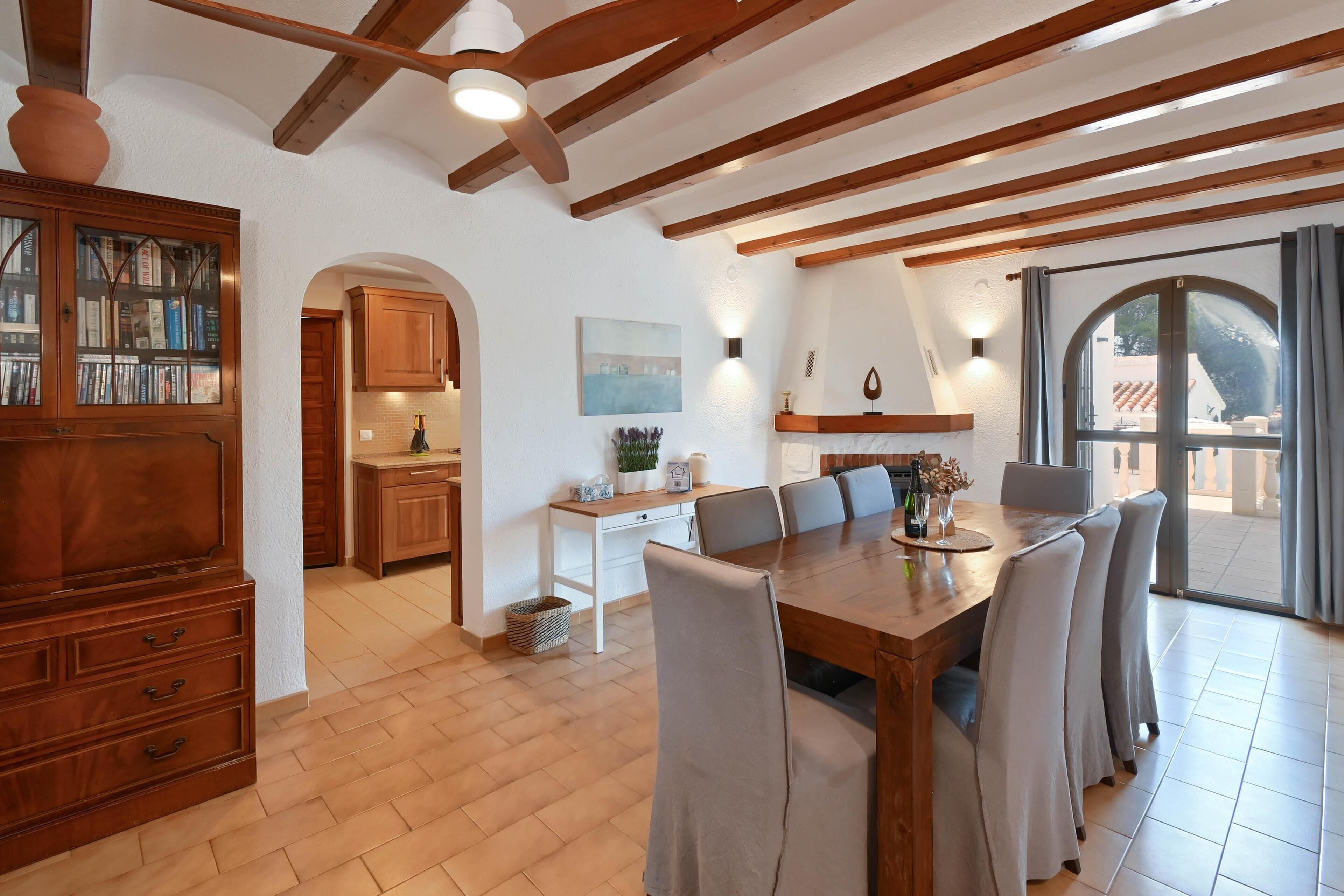 A dining area with a wooden table and chairs, a kitchen with wooden cabinets, and a view through a large window.