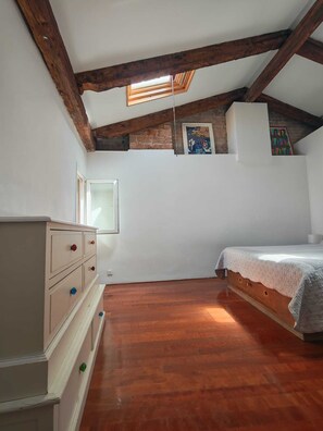 Une chambre avec un plafond en pente, des poutres en bois, un lit, une commode et une fenêtre.