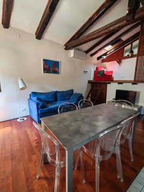 Une pièce avec un canapé bleu, une table à manger entourée de chaises transparentes, des poutres en bois au plafond et un tableau accroché au mur.