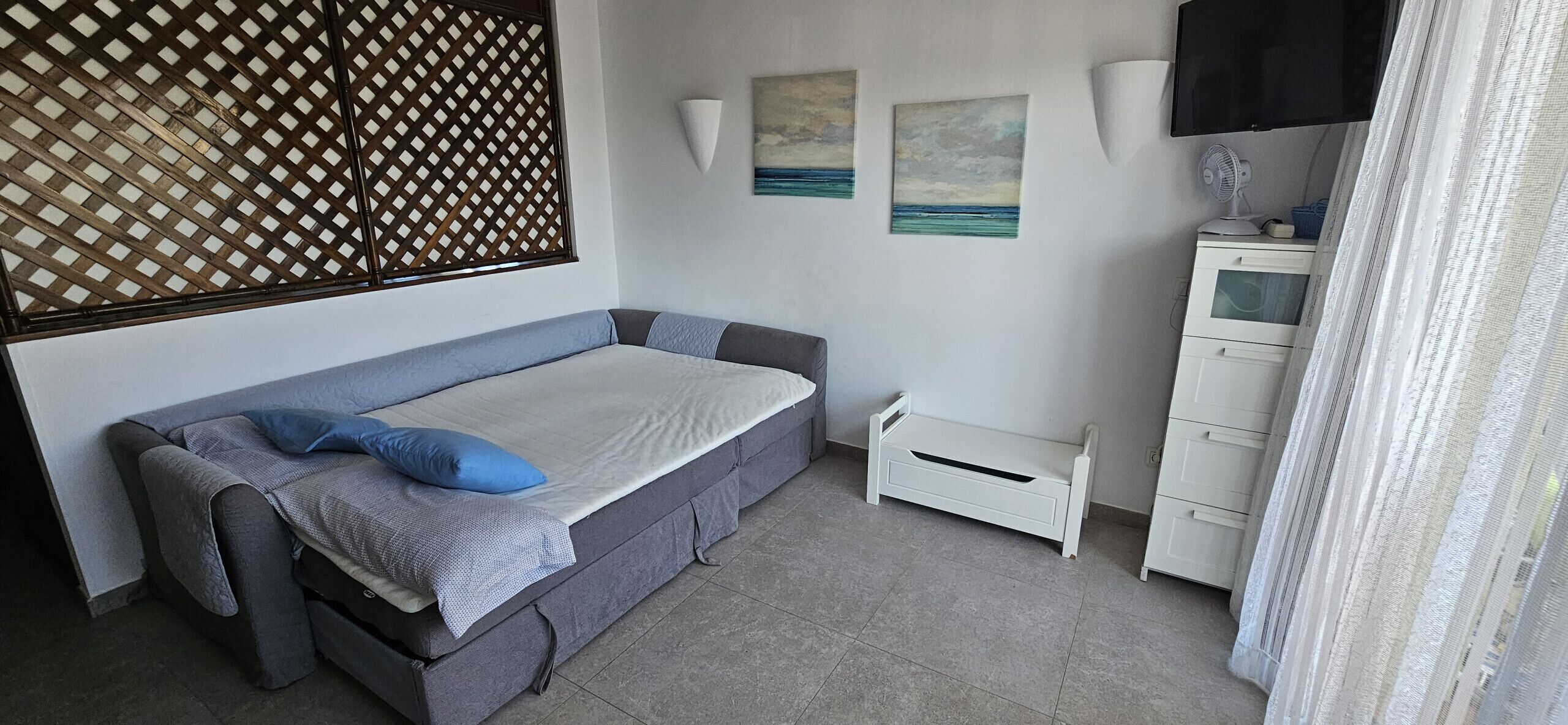 A room with a grey sofa bed, a small white bedside table, a wall-mounted lamp, and two paintings.