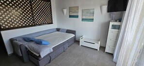 Um quarto com uma cama-sofá cinza, uma pequena mesa de cabeceira branca, uma luminária de parede e duas pinturas.