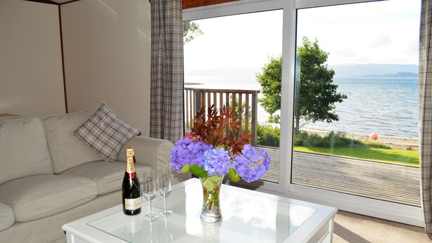 A room with a glass-top table holding a bottle of champagne, two glasses, and a vase with flowers. A white sofa with a plaid pillow is visible. A large window offers a view of a lake and greenery. - Cockleshell Lodge located at the waters edge near Otter Ferry-Pet friendly (Tighnabruaich)