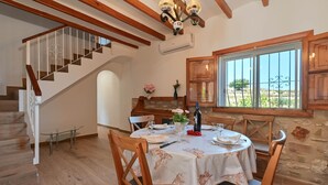 A dining area with a round table set for four, wooden chairs, a chandelier, a staircase, and a view through a window. - Rustic and cheerful holiday home in Javea, Costa Blanca, Spain  with private pool for 6 persons (Javea)