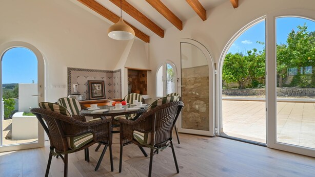 A dining area with a round table and four wicker chairs, large arched windows, and a view of the outdoors. - Rustic and cheerful holiday home in Javea, Costa Blanca, Spain  with private pool for 6 persons (Javea)