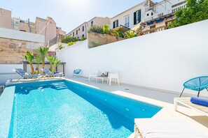 A swimming pool with lounge chairs and a table, surrounded by a white wall and buildings. - VILLA OASIS TERRA BLANCA (Palma de Mallorca)