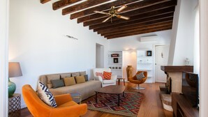 Ein modernes Wohnzimmer mit einer Couch, einem orangefarbenen Sessel und einem Couchtisch. Es gibt einen Kamin, Holzbalken an der Decke und einen Fernseher auf einem Holzgestell.