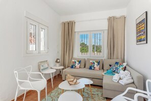 Een moderne woonkamer met een bank, twee witte stoelen, een bijzettafeltje en een raam met gordijnen.