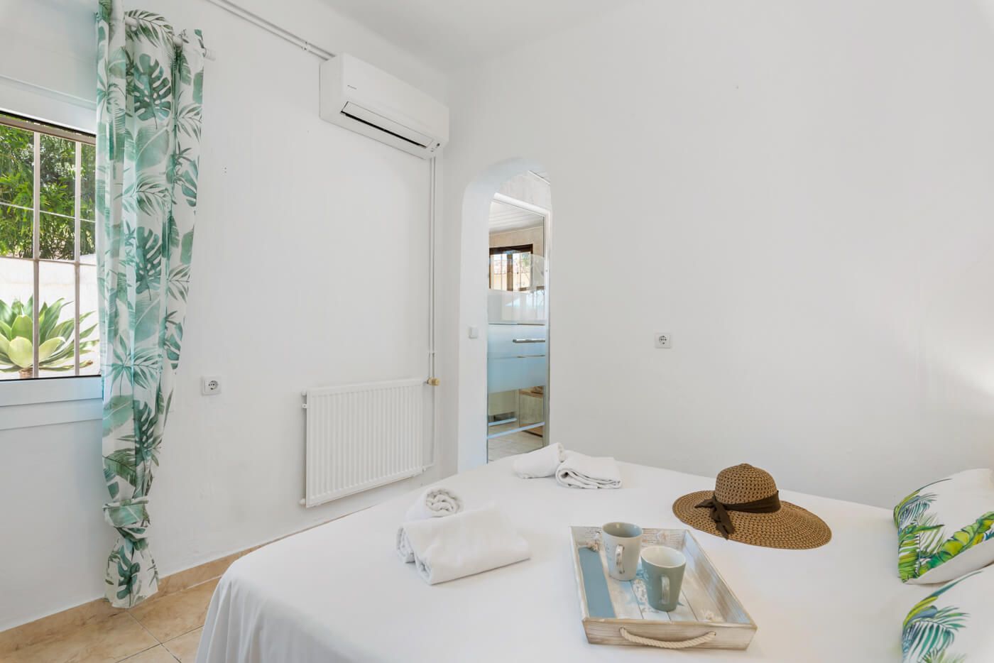 A bedroom with a bed, a radiator, a window with curtains, a bedside table with a tray, and a hat.