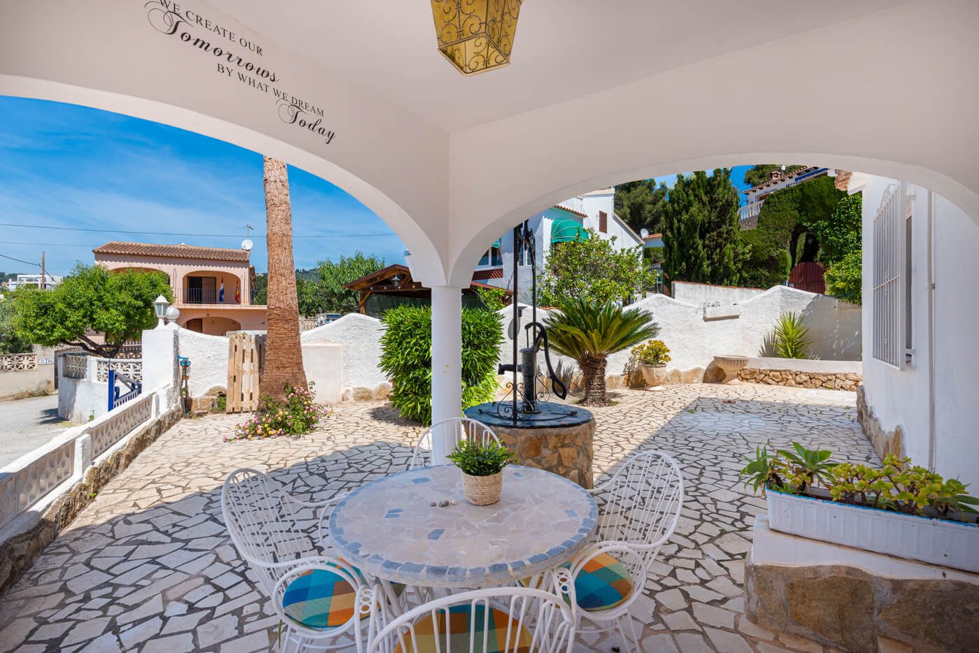 A covered outdoor patio with a round table and chairs, surrounded by stone flooring and potted plants.