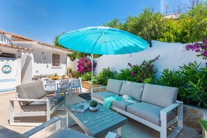 Een terras buiten met een blauwe parasol, een bank, stoelen en een eettafel.