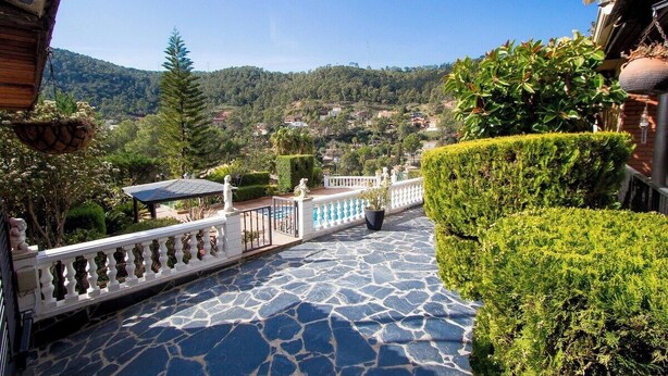 A stone-paved terrace with a white balustrade, lush greenery, and a view of a hillside village.