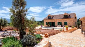 Une maison en pierre avec un toit en tuiles, une piscine et un jardin agrémenté de diverses plantes et d’un grand arbre.