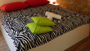 A bedroom with a zebra-patterned bedspread, red and green pillows, a painting of boats on water, a stone wall, and a nightstand with a lamp.