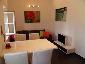 Un salon moderne avec une table à manger blanche, un canapé agrémenté de coussins colorés, un téléviseur à écran plat et des œuvres d’art accrochées aux murs.