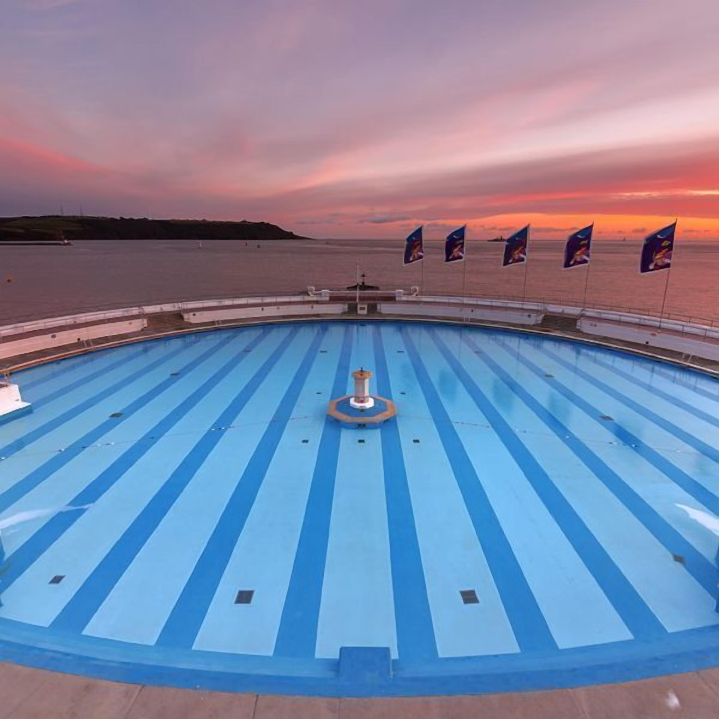 A pool deck with blue stripes and a central fountain, surrounded by flags and a sunset view.
