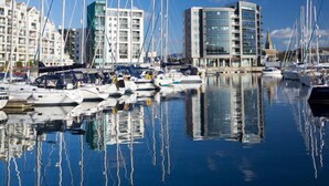 Een jachthaven met verschillende zeilboten afgemeerd, omringd door moderne gebouwen met glazen gevels.