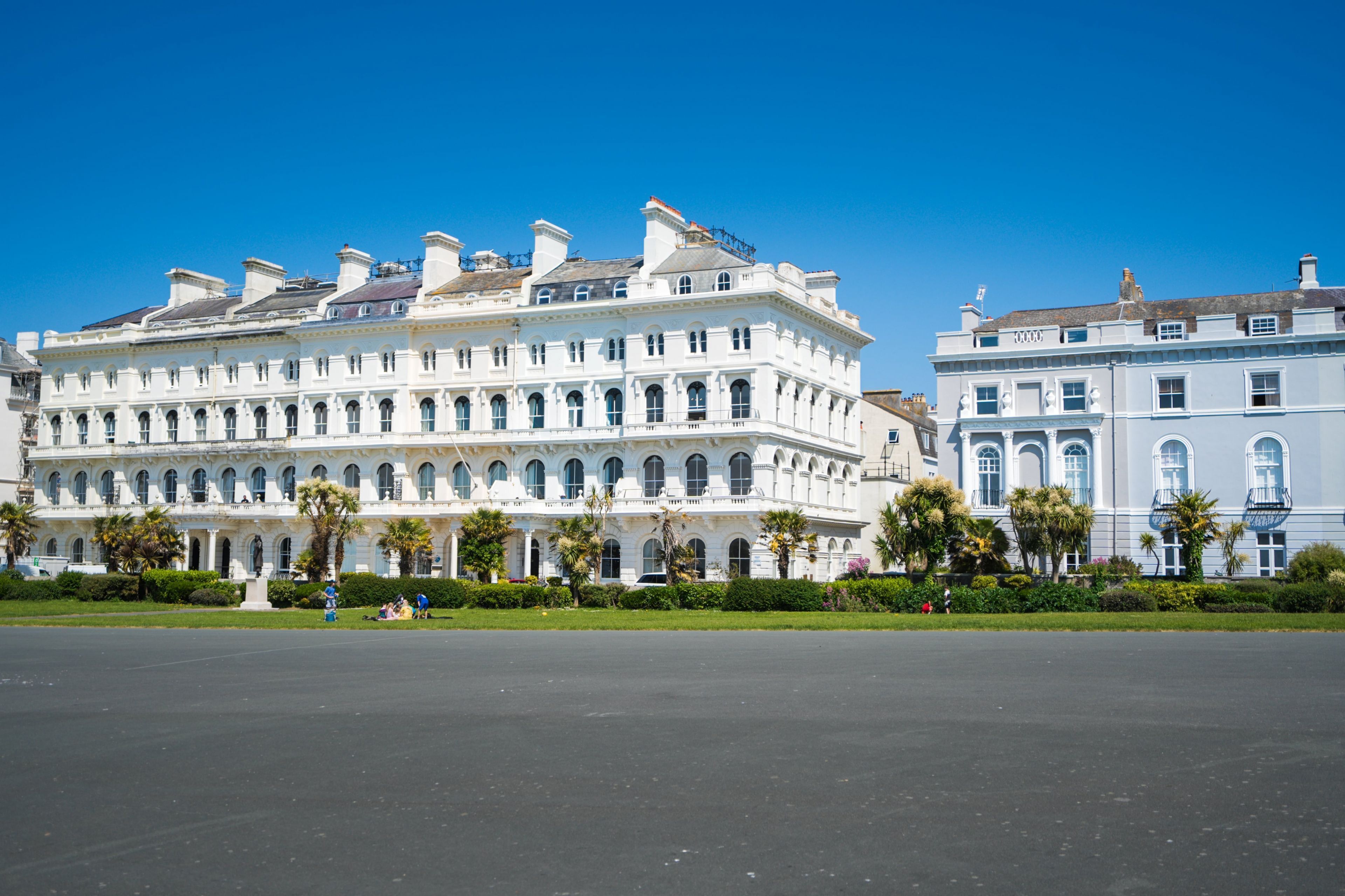 Un edificio grande e histórico con varios pisos y ventanas arqueadas, rodeado por un césped cuidado y palmeras.