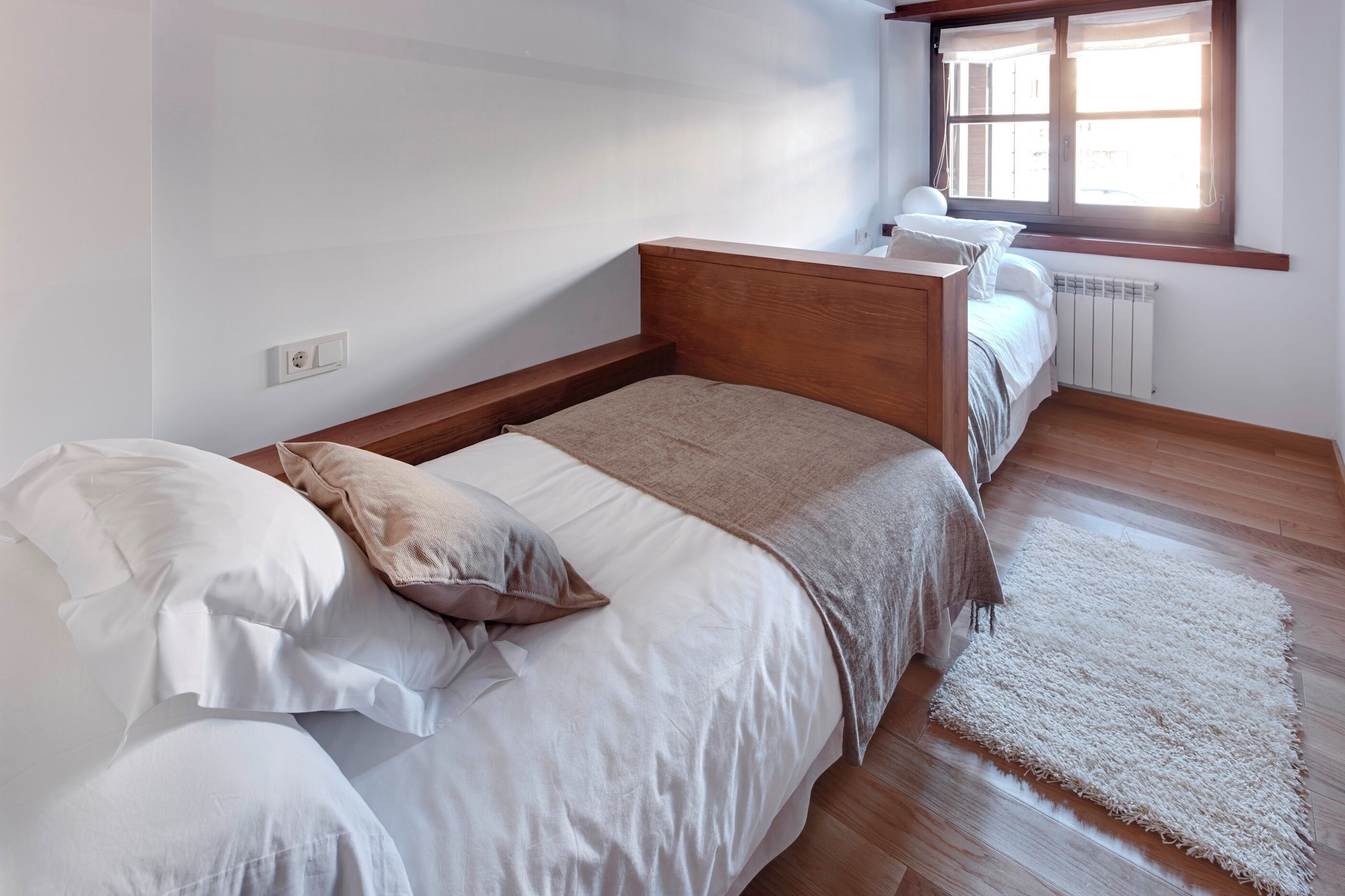 A bedroom with a wooden bed, white bedding, a white rug, and a window with curtains.