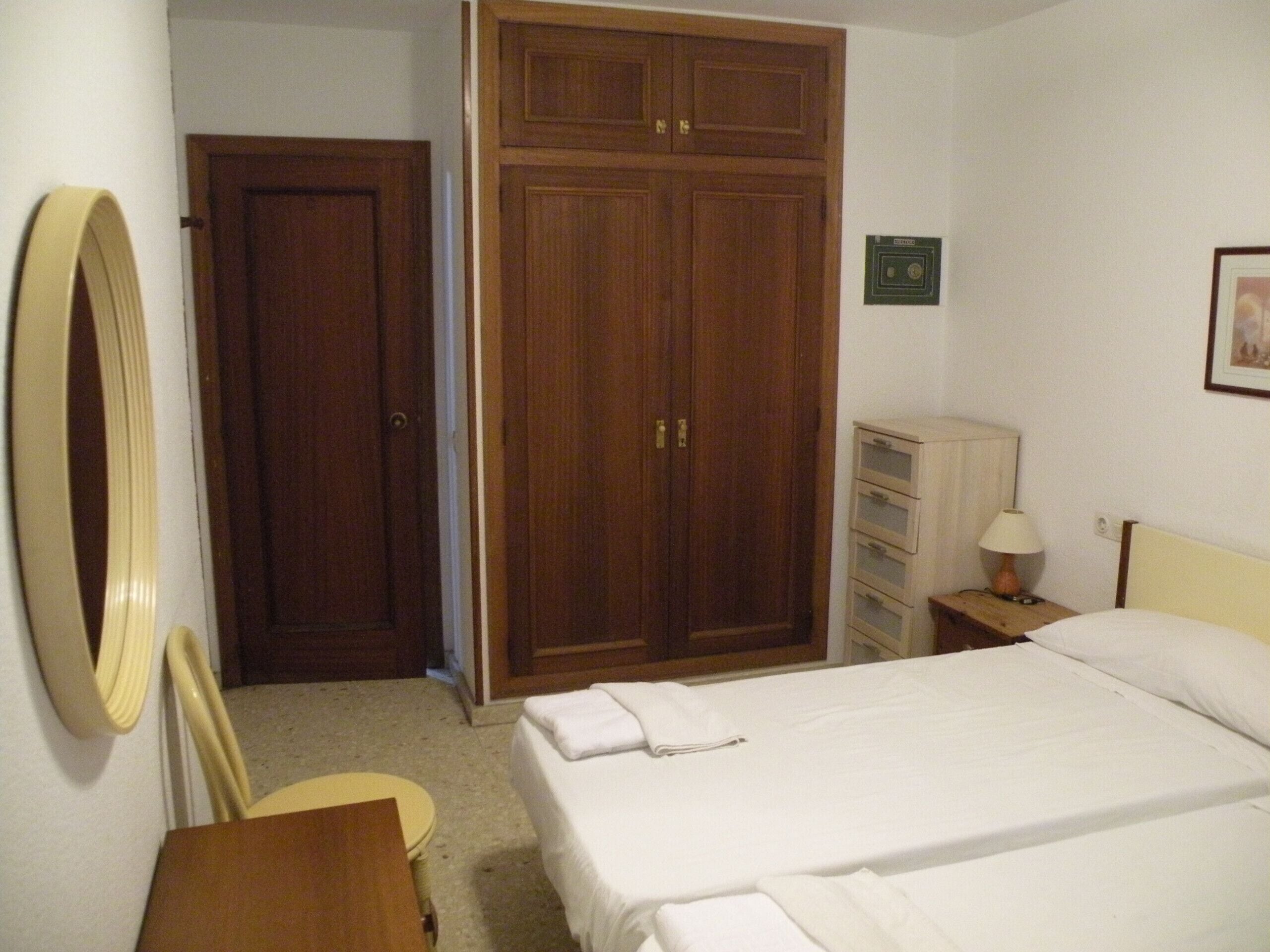 Habitación de hotel con cama, armario de madera, espejo, mesita de noche con lámpara y una silla pequeña.