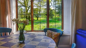 A room with a large window, a table with a checkered tablecloth, two chairs, a vase with flowers, and a view of a green landscape.