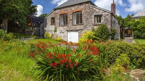 A stone house with a white door, surrounded by lush greenery and red flowers.