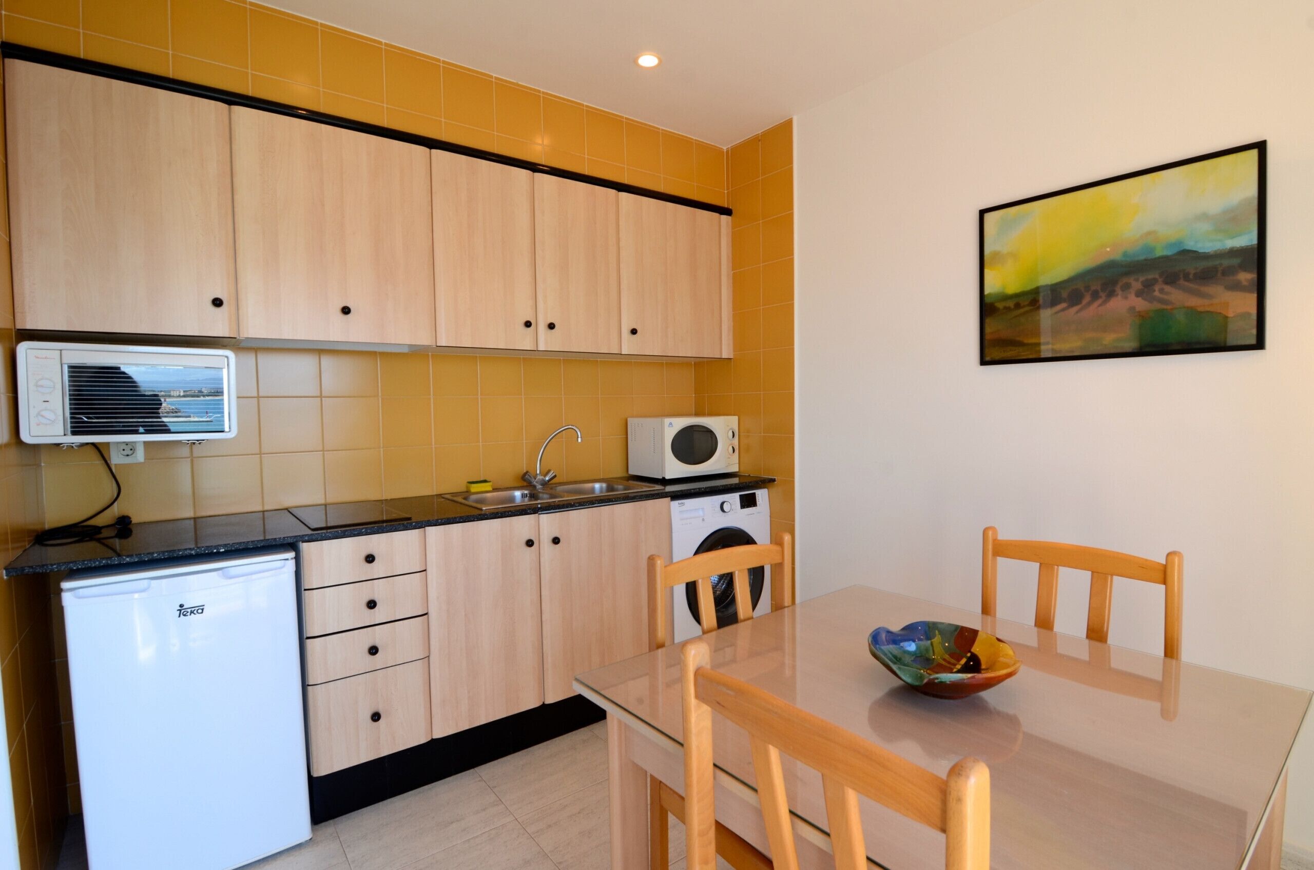 Une cuisine compacte avec des meubles de couleur bois clair, un plan de travail noir, un petit réfrigérateur, un four à micro-ondes et un lave-linge. On y trouve une table à manger avec des chaises et un saladier posé dessus.