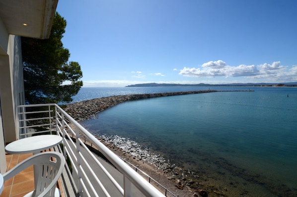 Un balcon offrant une vue sur une mer calme, un brise-lames rocheux et un ciel dégagé.