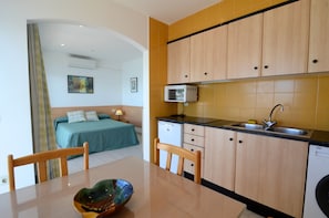 A compact kitchen with wooden cabinets, a sink, and a dishwasher. A bed with a green blanket is visible in the adjacent room.