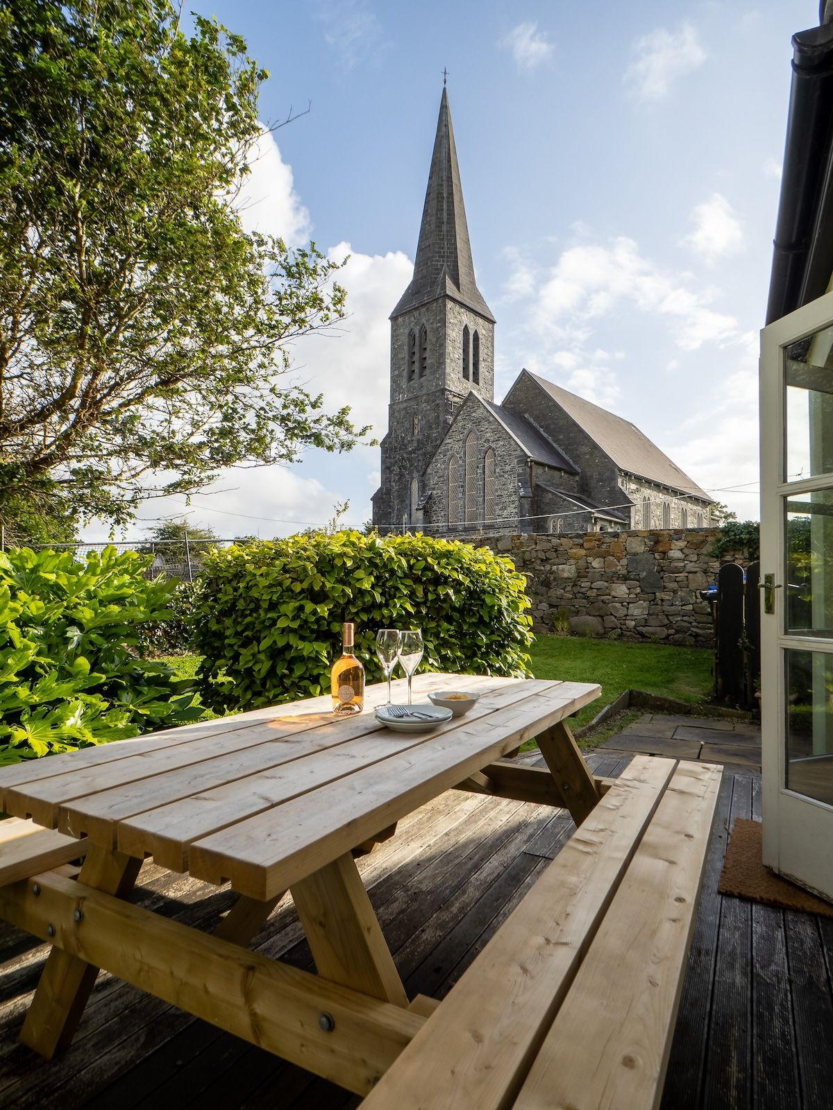 A wooden picnic table with a bottle and glasses, set against a stone wall and a church with a tall steeple.