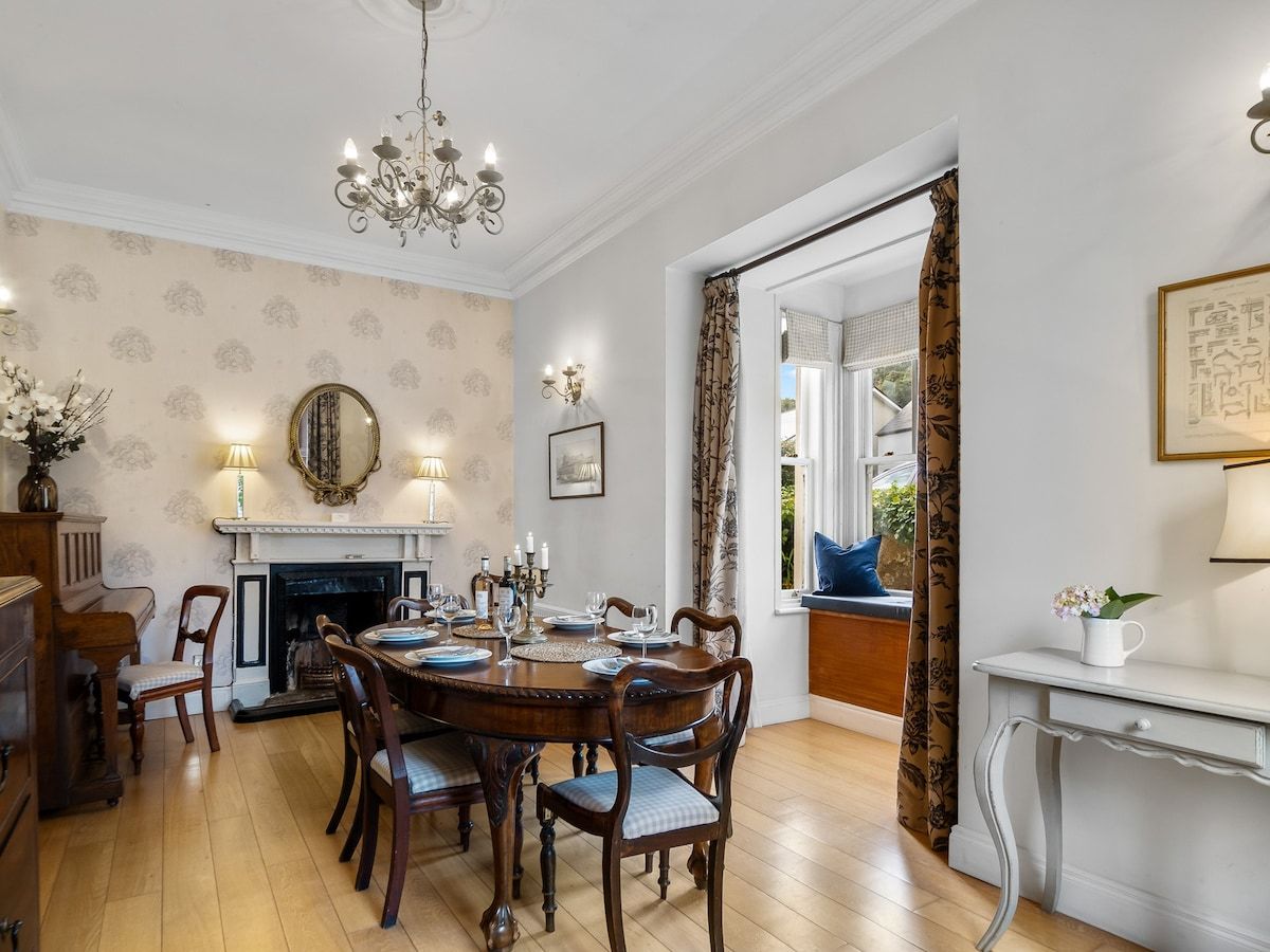 A dining room with a round table, chairs, a chandelier, a fireplace, and a view through a window.